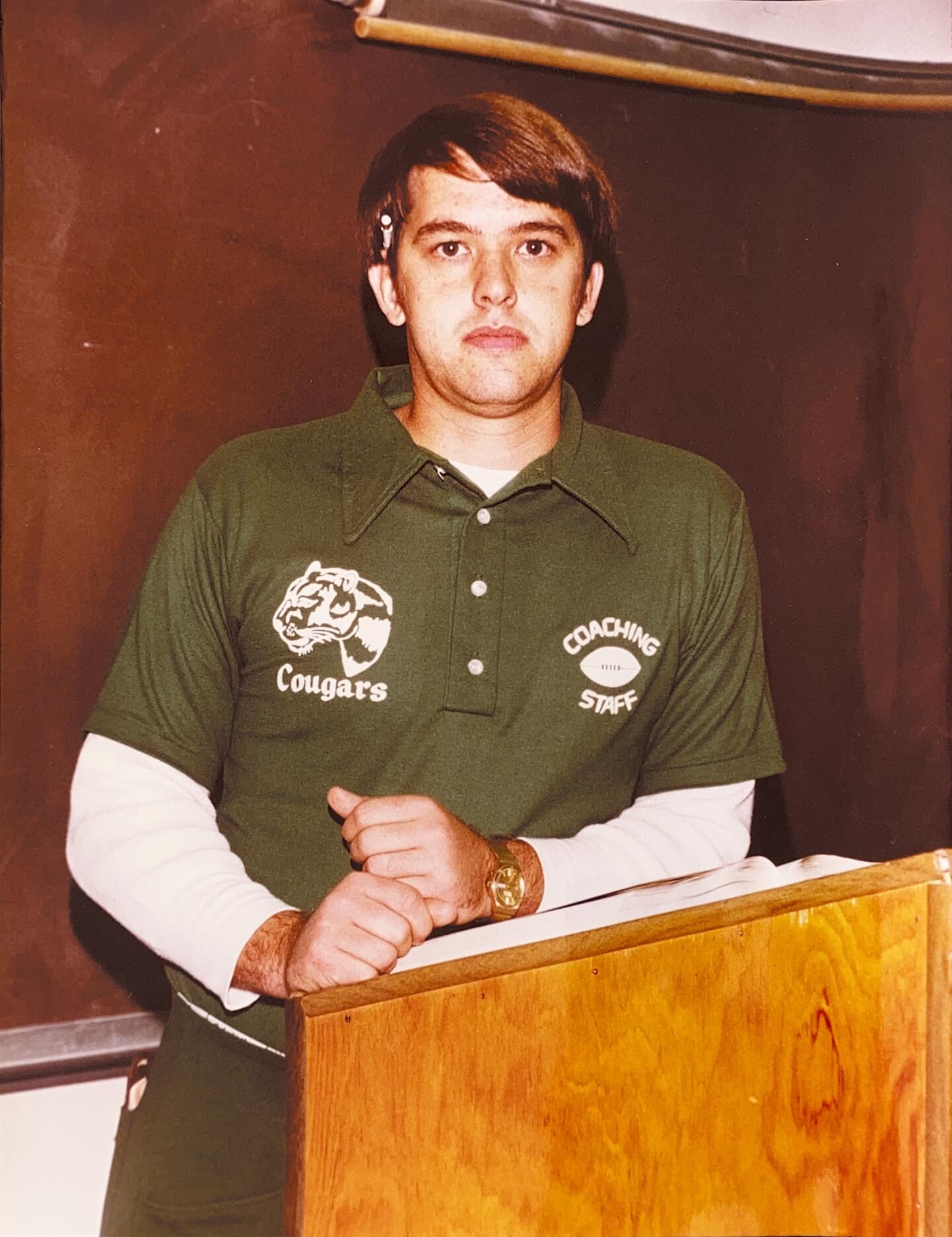 A young John Stallings, wearing a green Mountain Heritage Cougars coaching staff polo shirt, stands behind a lectern.